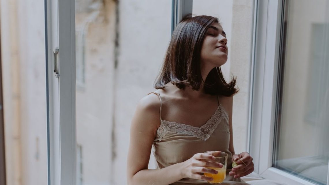 Woman in a spaghetti strap dress savoring orange juice on a window sill, indoors.