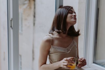 Woman in a spaghetti strap dress savoring orange juice on a window sill, indoors.