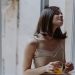 Woman in a spaghetti strap dress savoring orange juice on a window sill, indoors.